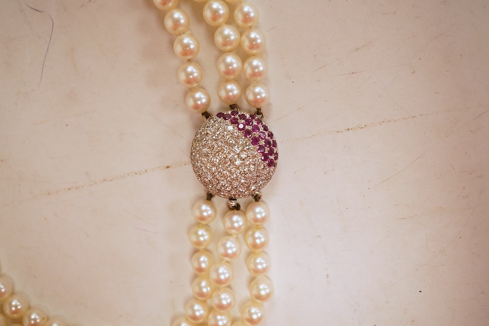 A cultured pearl, ruby and diamond clasp, composed of three rows of cultured pearls measuring approximately 6.9mm diameter, to a white precious metal (tests as 14k) disc-shaped clasp pavé-set with brilliant-cut diamonds